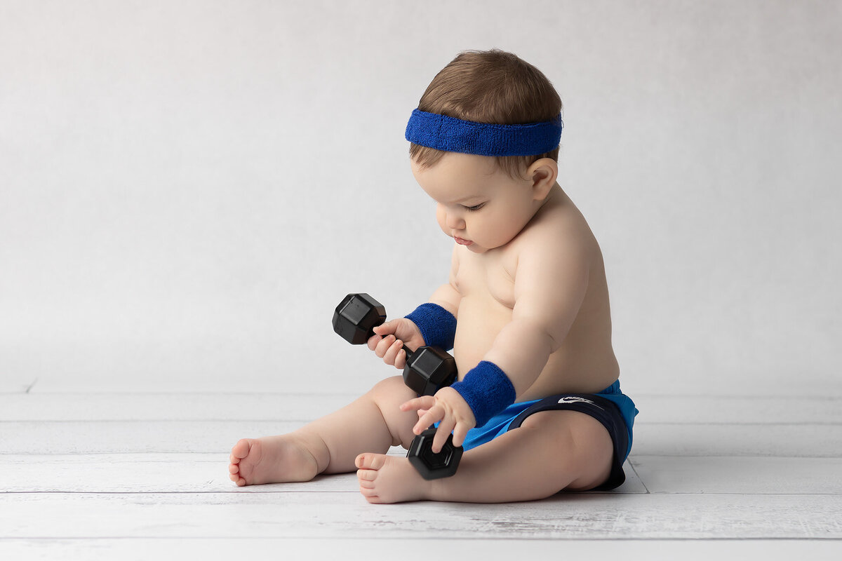 Baby wearing blue shorts, sweatbands, and a headband sits on a white wood floor while curiously examining two small dumbbells, captured by a Dallas milestone photographer.
