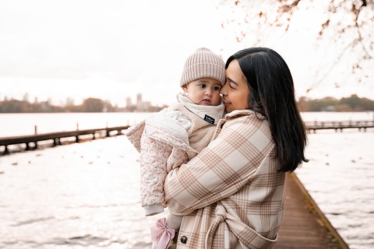 Mother cuddling baby by lake – Woman in a plaid coat lovingly holding her bundled baby near calm waters and a wooden pier on an overcast autumn day.