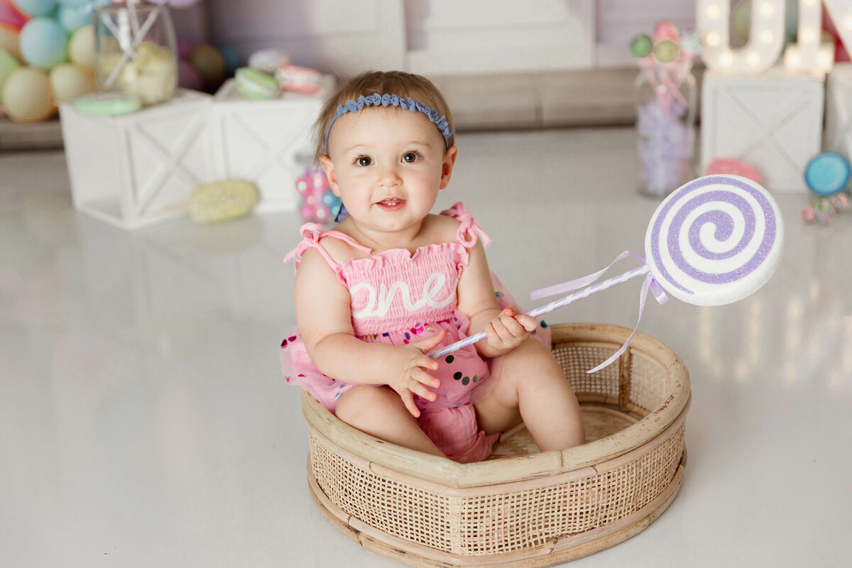 sweet one cake smash session in harrisburg pa baby girl in a pink onesie and a blue bow holding a purple swirly lollipop