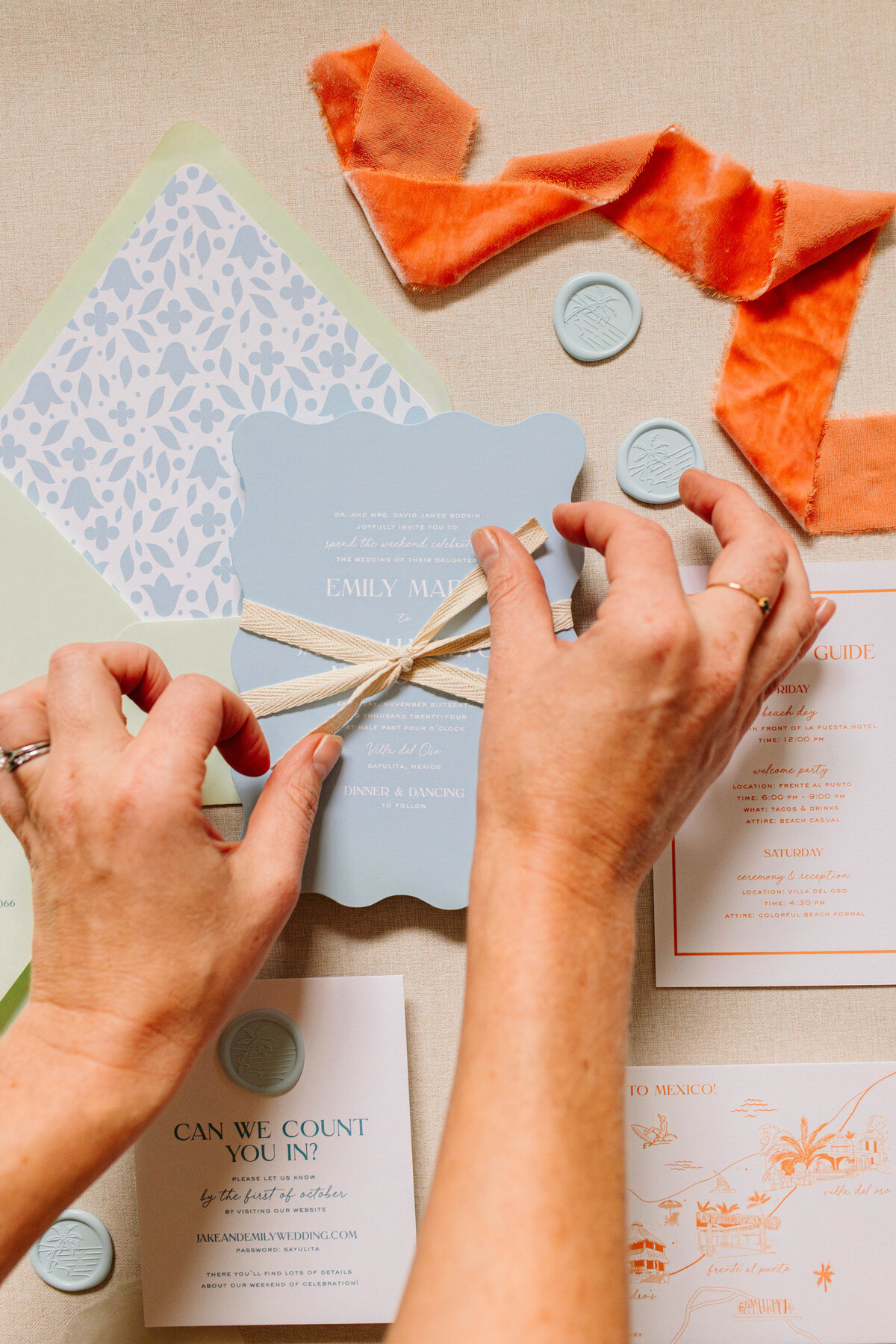 A pair of hands ties a ribbon around a scalloped blue wedding invitation, surrounded by coordinating stationery, wax seals, and an orange velvet ribbon. The elegant suite features soft pastel colors and intricate details.