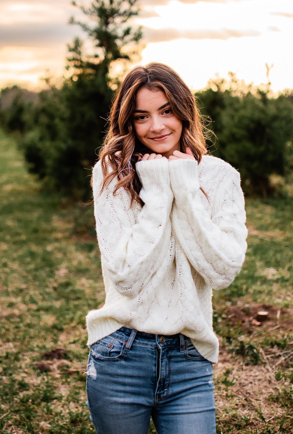 A high school senior wearing a white sweater poses at a Christmas tree farm in Manvel, TX.