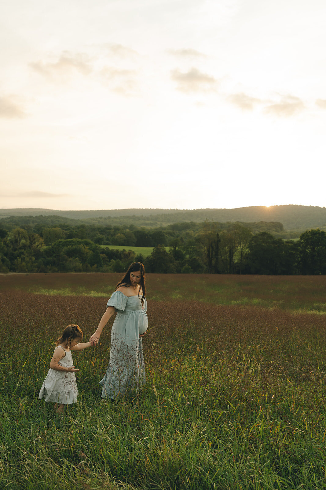 Pregnant mother walking hand-in-hand with her daughter through a sunlit field during a candid outdoor maternity session
