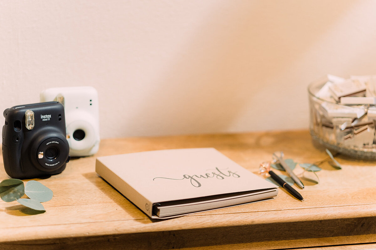 A wooden table setup featuring a guestbook labeled "guests" with two instant cameras, pens, and a glass bowl of snacks or mementos for guests.