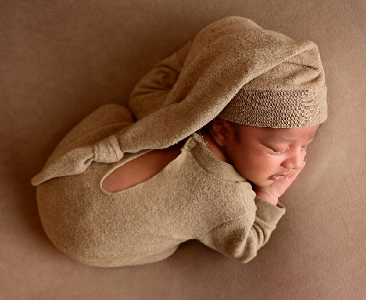 A newborn sleeping on his belly on a tan blanket wearing a tan outfit and  hat photographed from above by E. Benecki Photography.