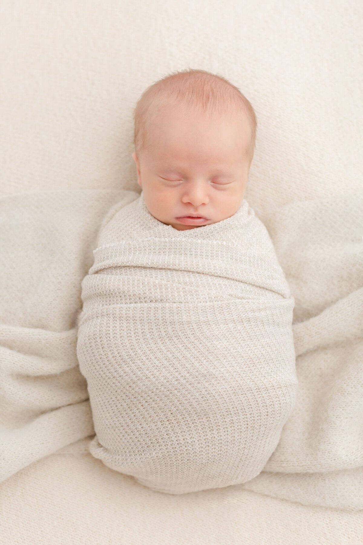 Light-skinned baby wrapped in beige textured cloth and sleeping on a cozy beige blanket. Baby is sleeping peacefully on his back.