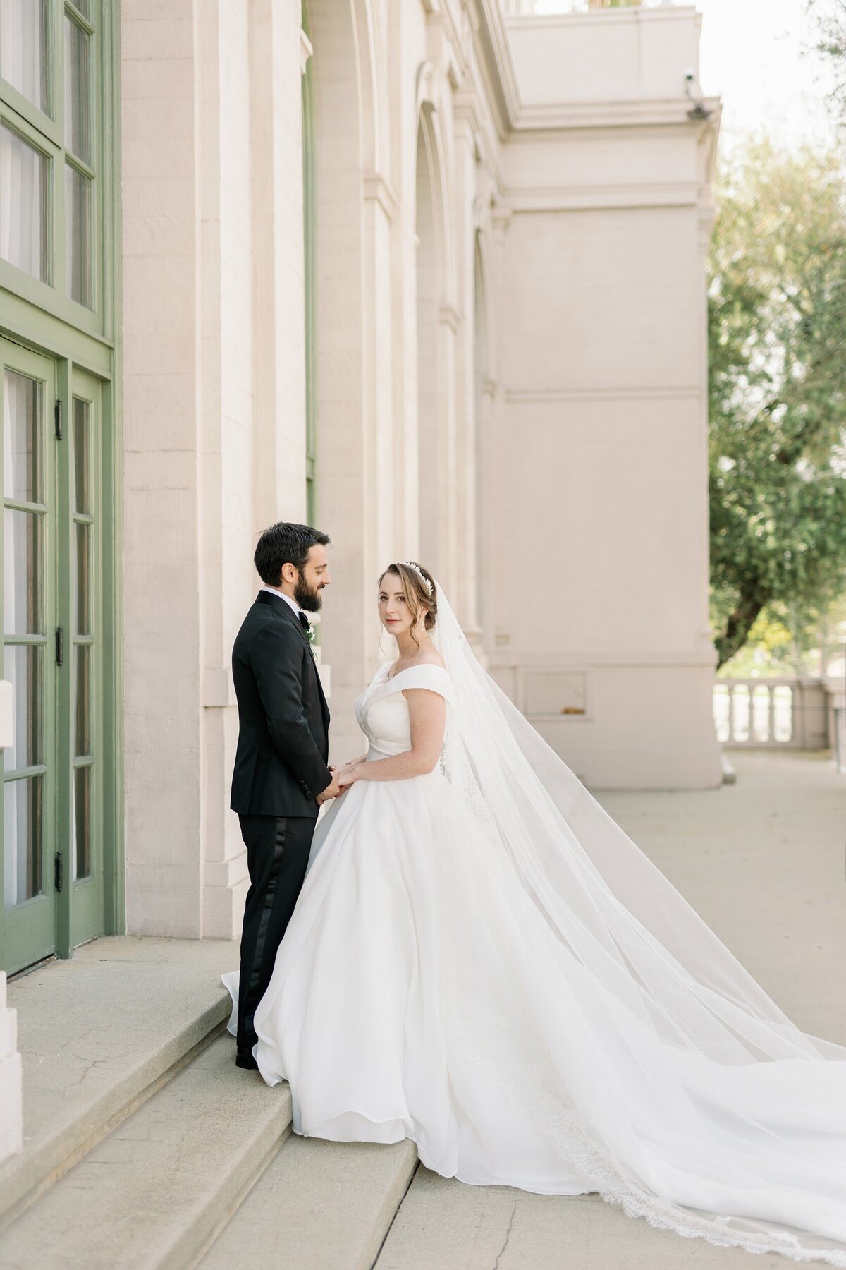 Classic couple portrait with bride in soft makeup and a half-updo wearing an off-shoulder ballgown and long veil on the steps of a historic estate portico.