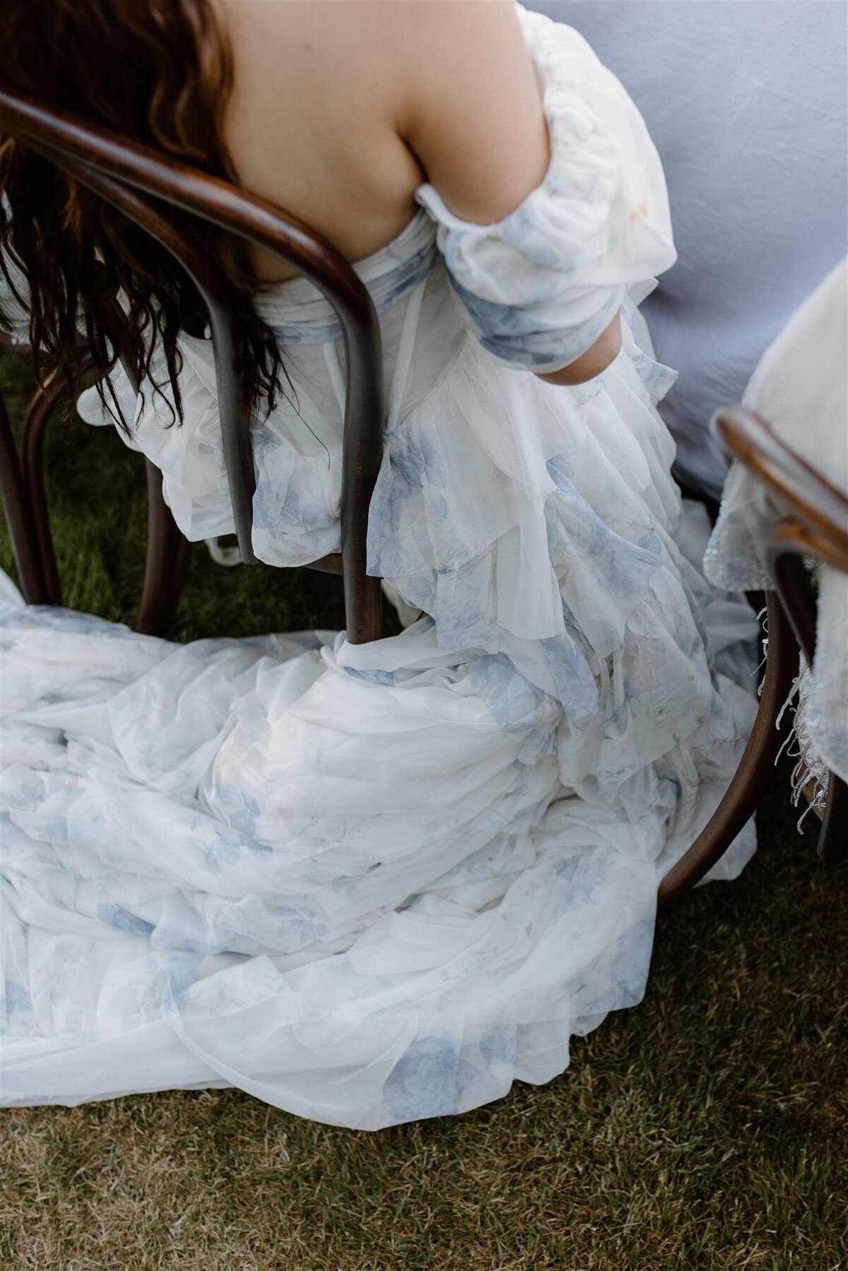 Close-up of a bride in an off-shoulder, blue-and-white floral gown sitting on a wooden chair, with the train of her ruffled dress flowing onto the grass.