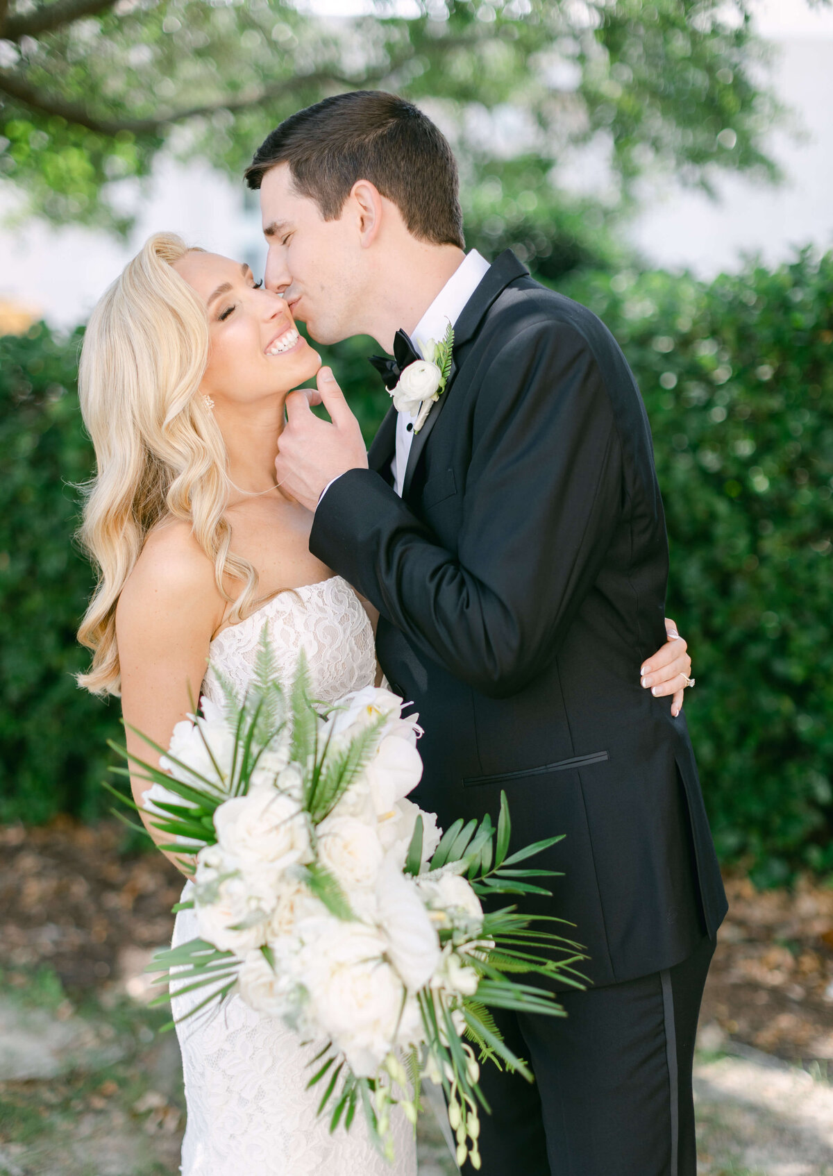 A bride smiles while a groom kisses her on cheek.