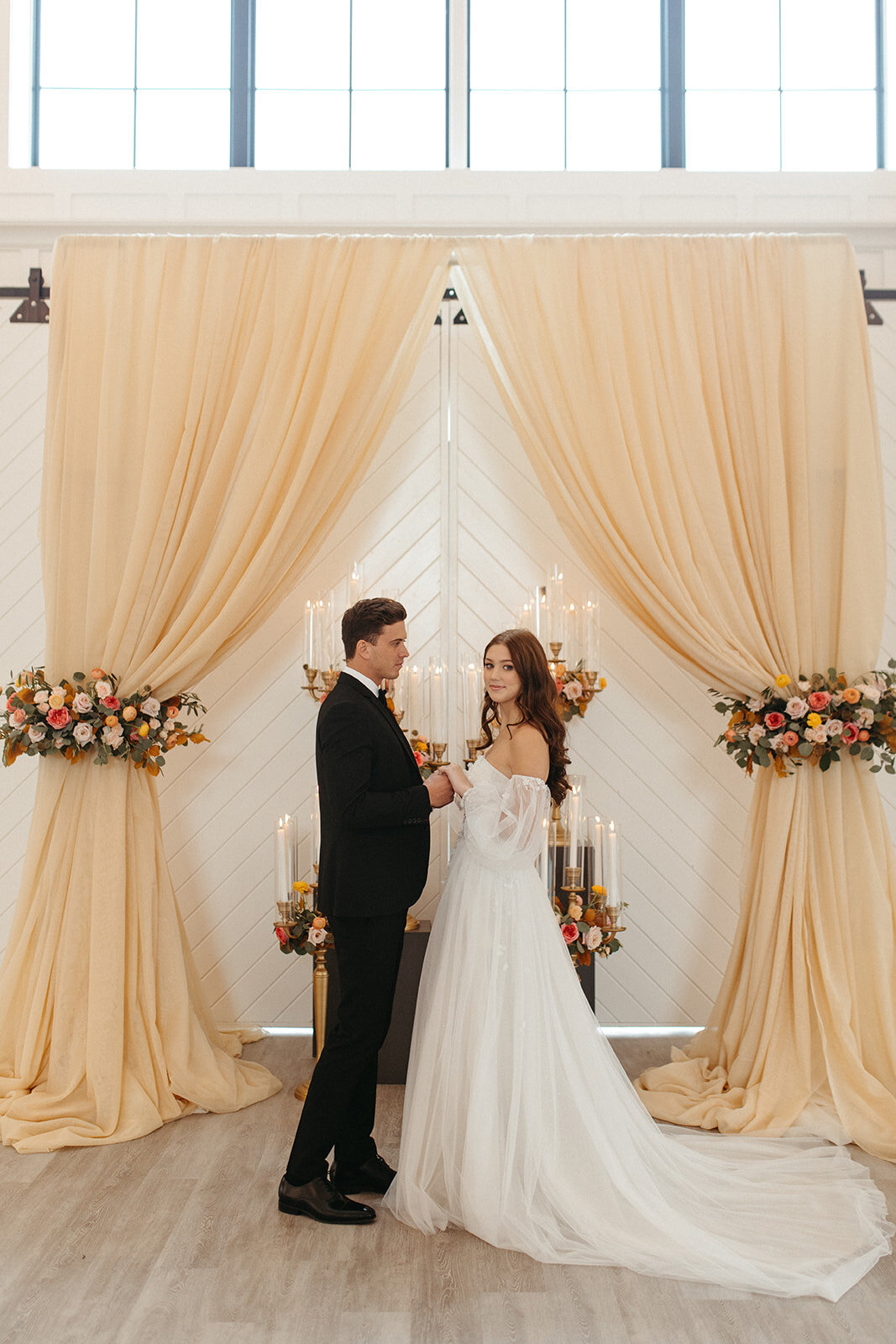 A bride and groom wearing a white wedding gown and black tuxedo hold hands in a bright and airy room filled with candles.