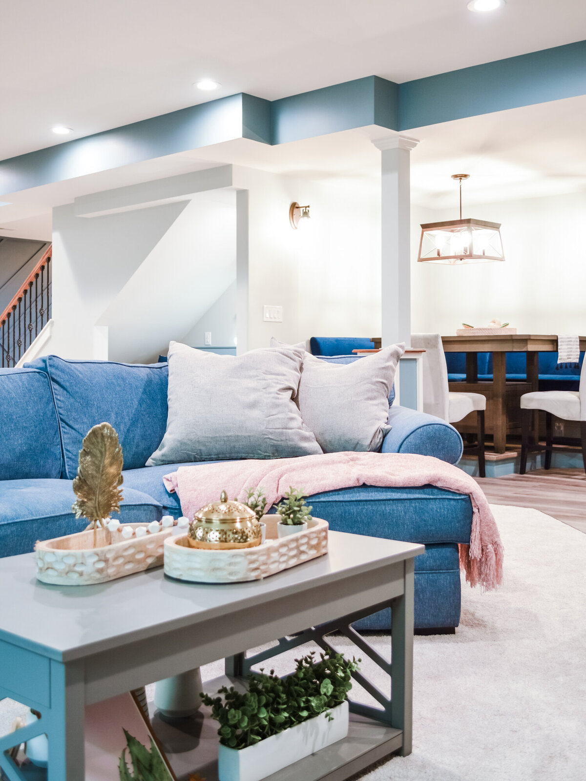 A spacious and modern living area with light gray walls and a recessed ceiling with a teal accent. A large blue sectional sofa with light gray throw pillows and a soft pink blanket occupies the left side of the frame. A light gray coffee table with a lower shelf holding plants sits in front of the sofa, displaying decorative trays and a glass cloche. In the background, a dining area with a light wood table and blue chairs is visible, illuminated by a modern pendant light. A staircase with dark railings is partially visible on the far left. The floor is covered with a light-colored rug in the living area and appears to be hardwood in the dining space. The room is well-lit by recessed ceiling lights and a wall sconce.
