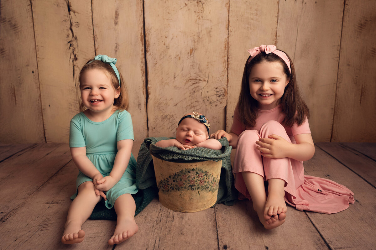 Portrait of newborn baby girl with two older sisters. Baby is asleep in a bucket with a sister sitting on either side.