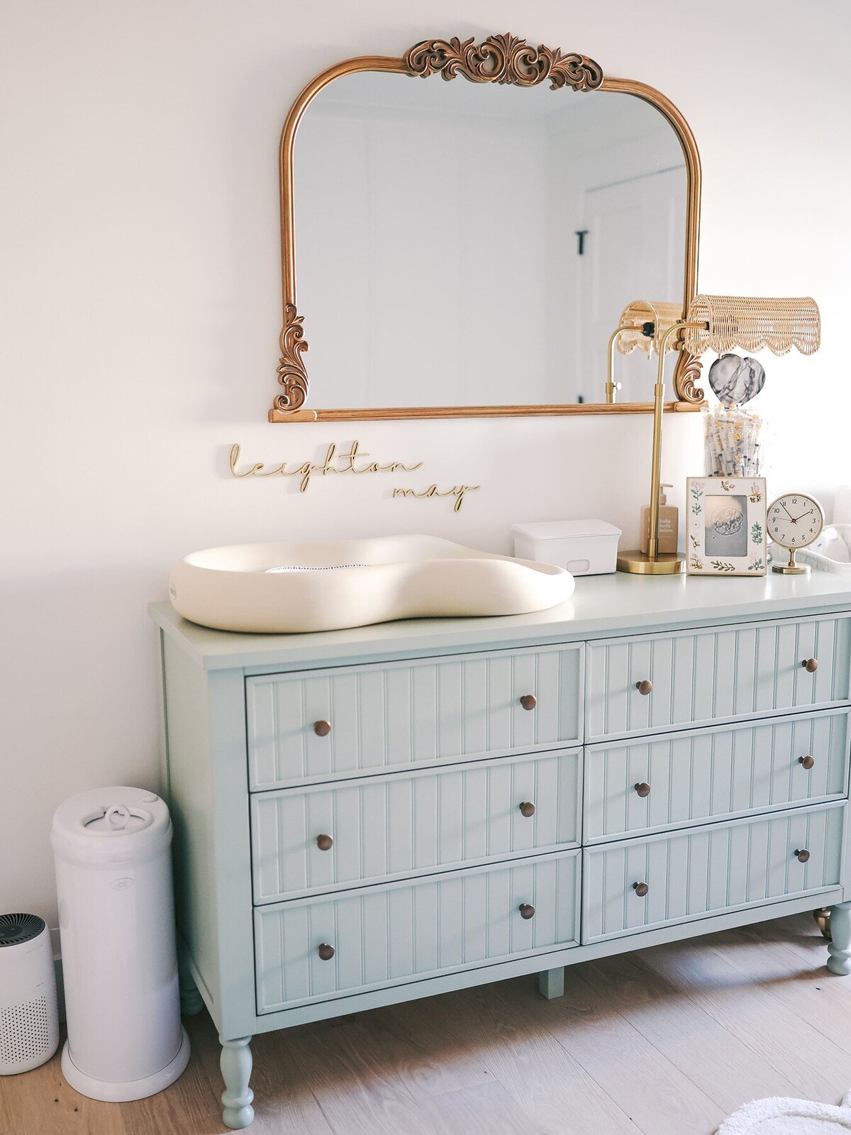 A dresser with picture frames, a clock, changing pad, and lamp with a mirror above it.  A nursery for a baby girl.