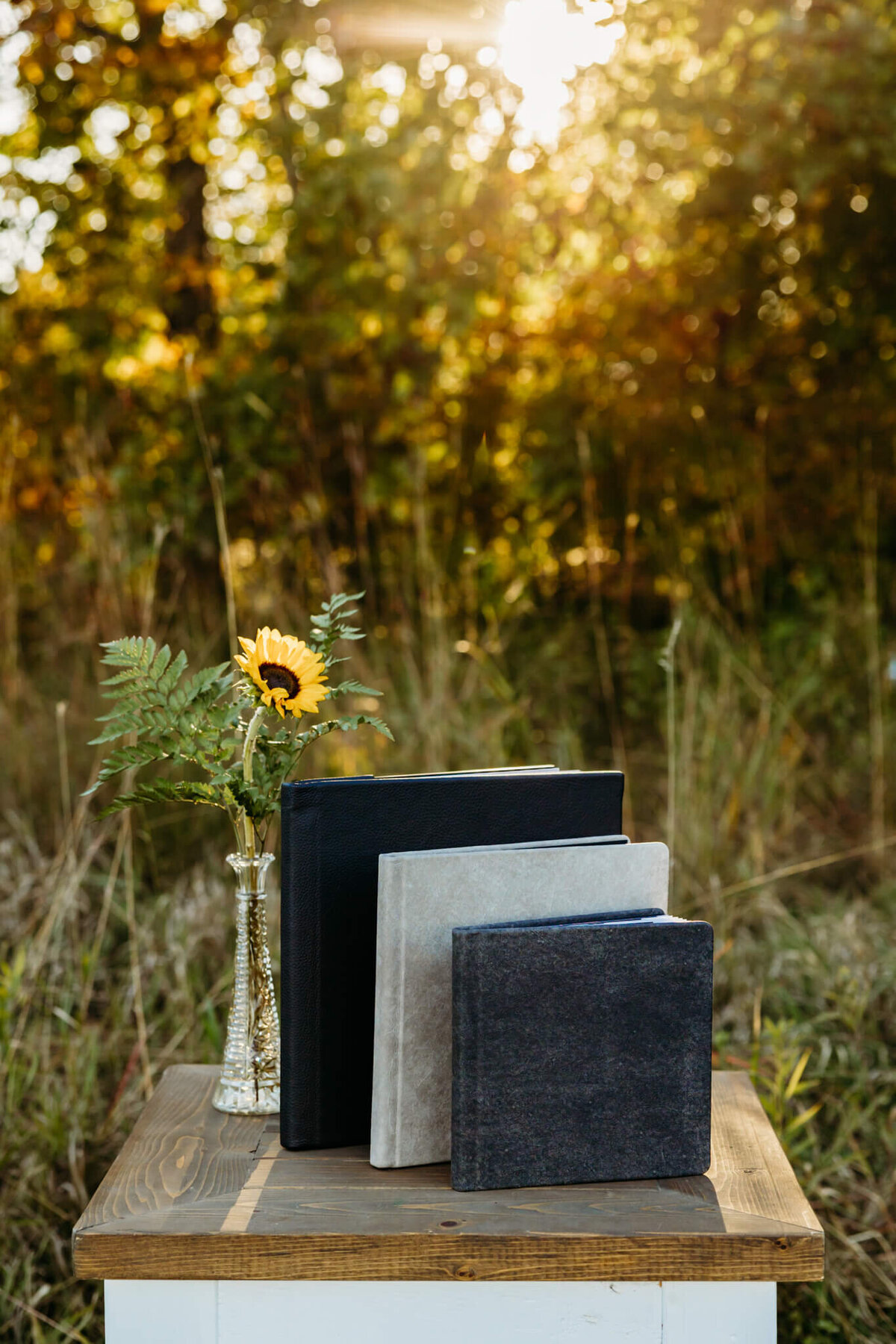 three neutral colored photo albums standing up on a wooden table by a sunflower as the sun sets behind them