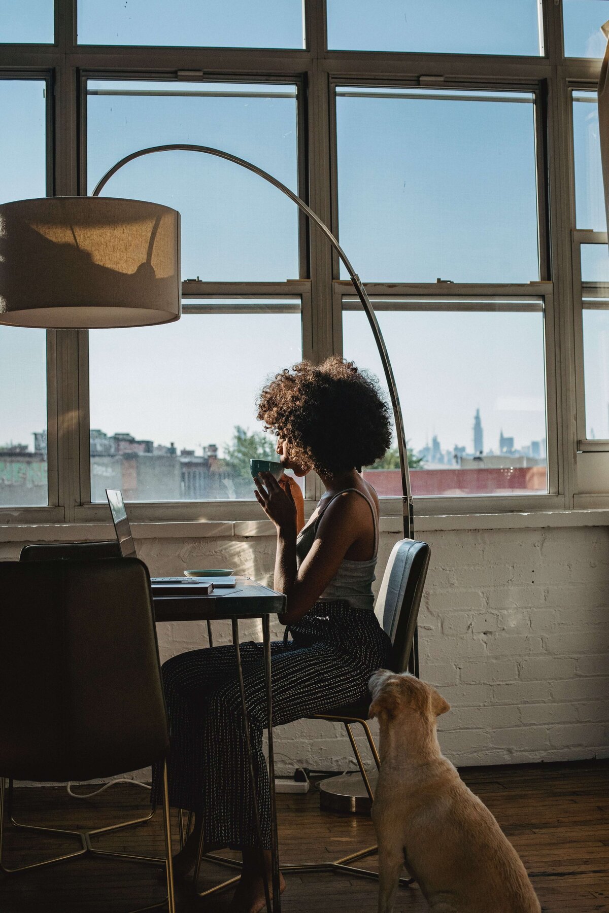 Woman sips coffee at a table in front of open windows while a dog sits by her chair
