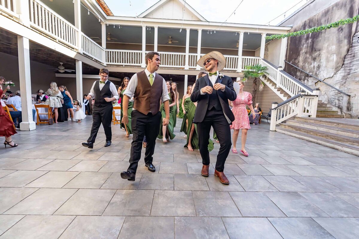 A group of people dressed in formal and semi-formal attire dancing enthusiastically outdoors on a tiled courtyard. One man wears a cowboy hat and boots while others join the fun in suits, dresses, and green gowns.