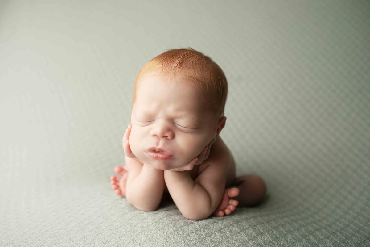 Red-headed newborn boy posed on a plush backdrop, his tiny hands resting under his chin in a sweet and serene moment