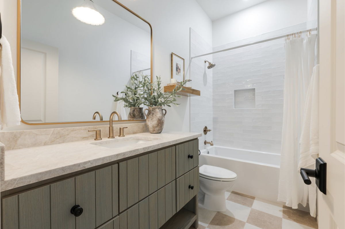 Guest bathroom with green stained vanity, brass fixtures, oversized mirror, and white subway-tiled tub surround. Neutral checkered floor tile and natural accents complete the space.