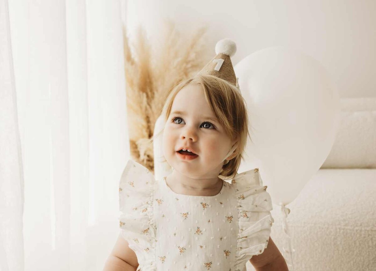 Ruua Hamdan Photography (UK): A baby in a floral dress and golden party hat looking up while smiling.