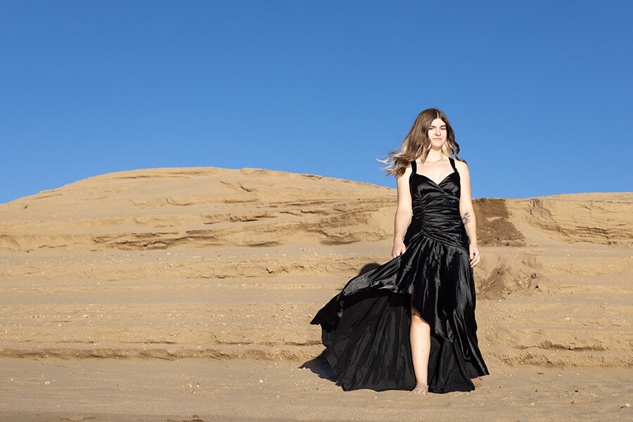 High school senior in long black dress walking over sand dune.