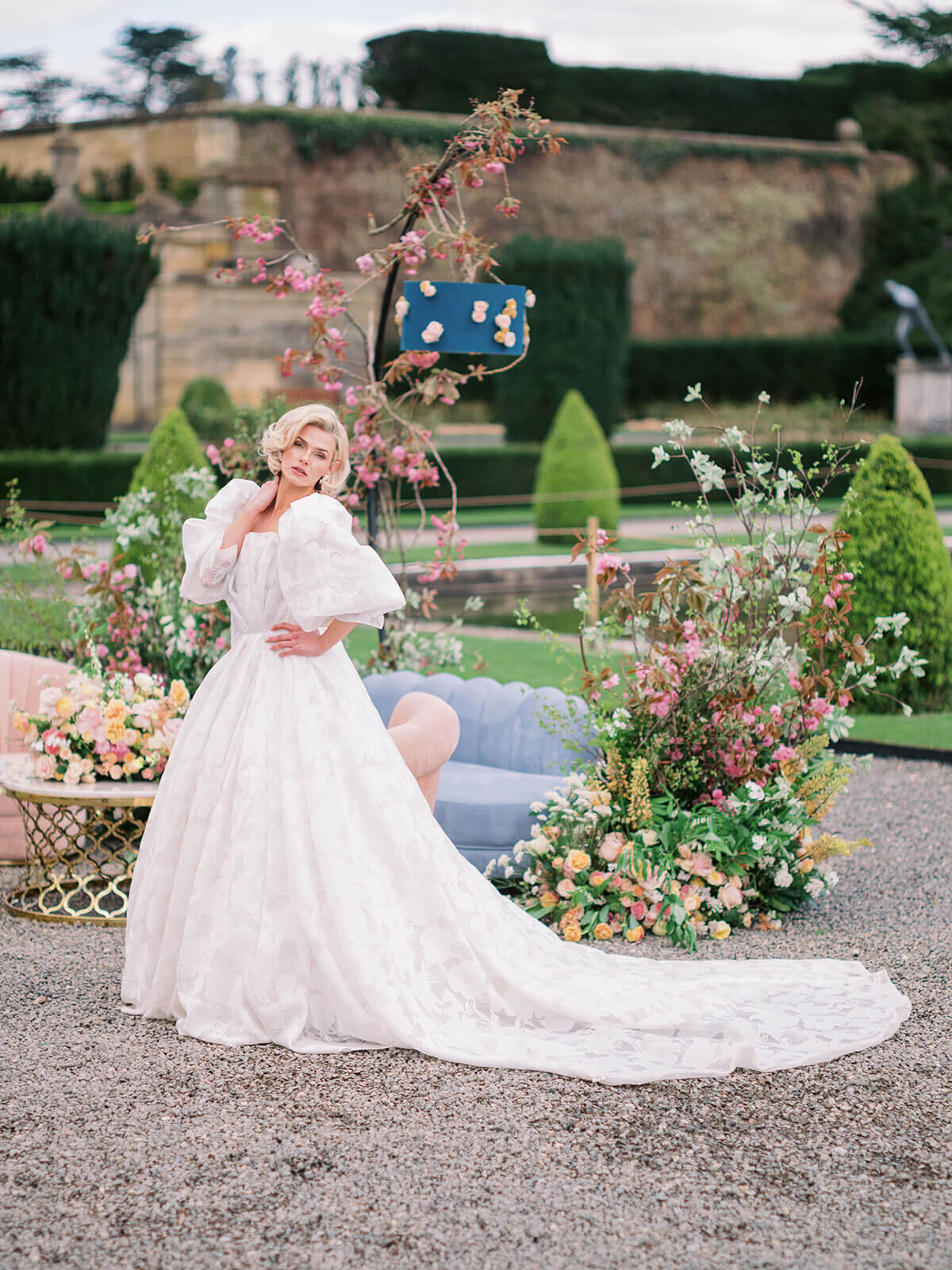 bride standing in front of a wedding drinks reception lounge set up in the gardens of blenheim palace with pink and blue seating and pink and peach flowers