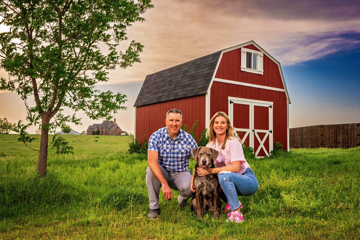 Dog mom and dad kneel in the grass with a silver lab in front of a red barn.
