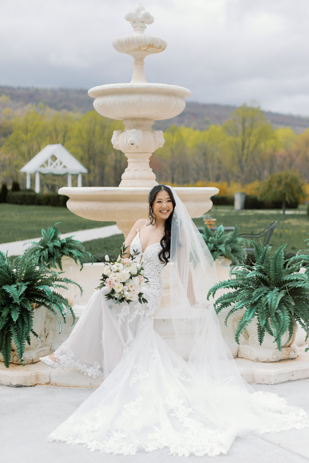 Bride in lace gown with cathedral veil posing beside stone fountain at Springfield Manor.