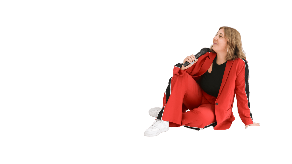 A person in a red suit and white sneakers sits on the floor, holding a microphone and smiling while looking to the side, against a plain white background.