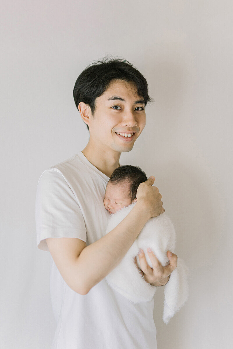 Father is holding baby in his arms close to his chest and smiling. Shot in Yokohama by newborn photographer Yukika Ishikawa Photography
