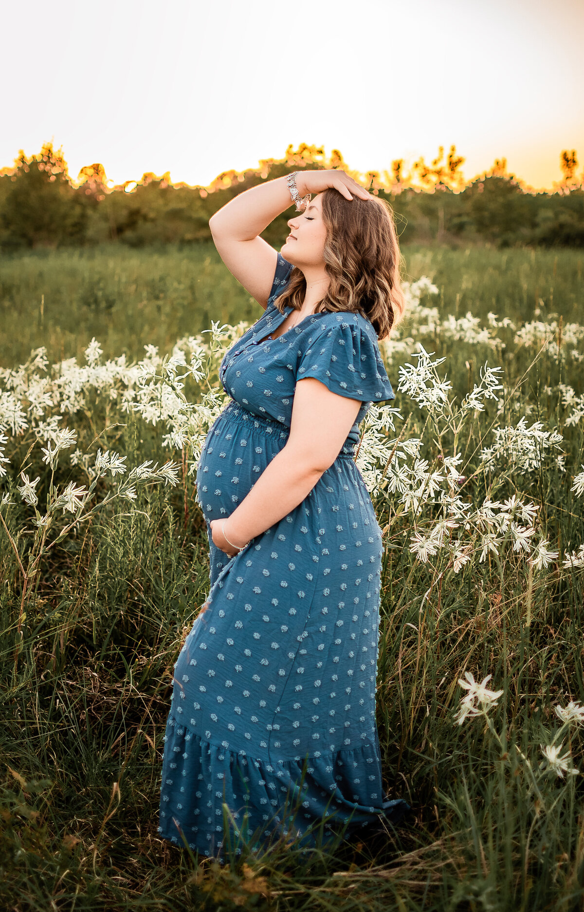 At a Harris county park, a mom to be stands in a field of white flowers as she looks toward the sky and holds her baby bump.