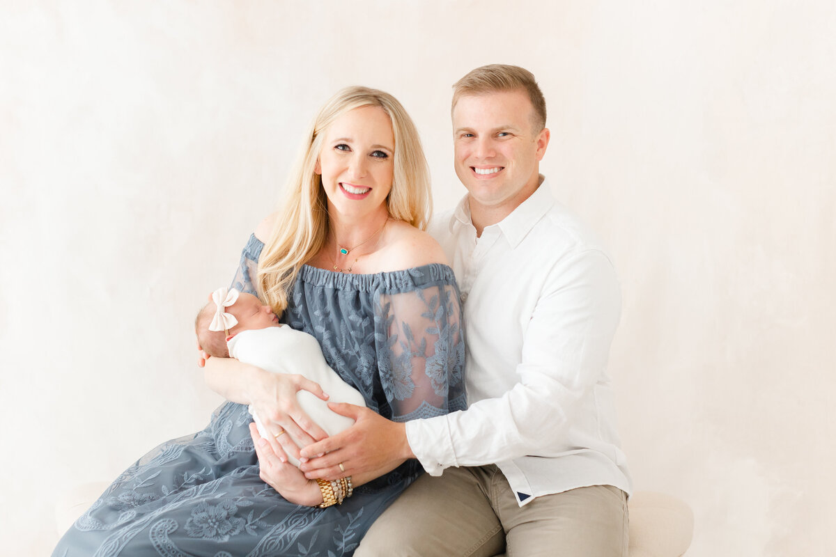 A DC newborn photography photo of a mother and father looking at the camera while sitting on a bench and holding their newborn child