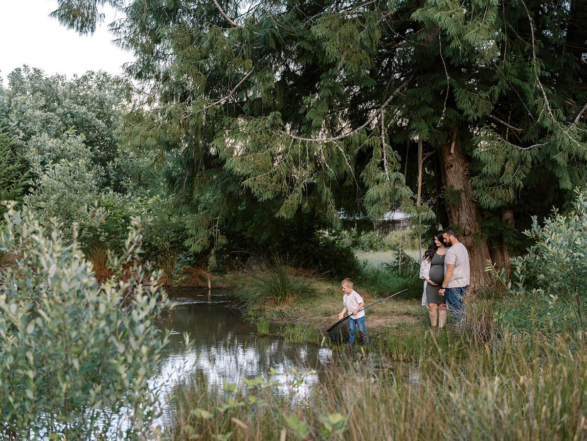 A couple watches their son fishing with a net next to a pond during a family photography shoot by Among the Oaks photography.