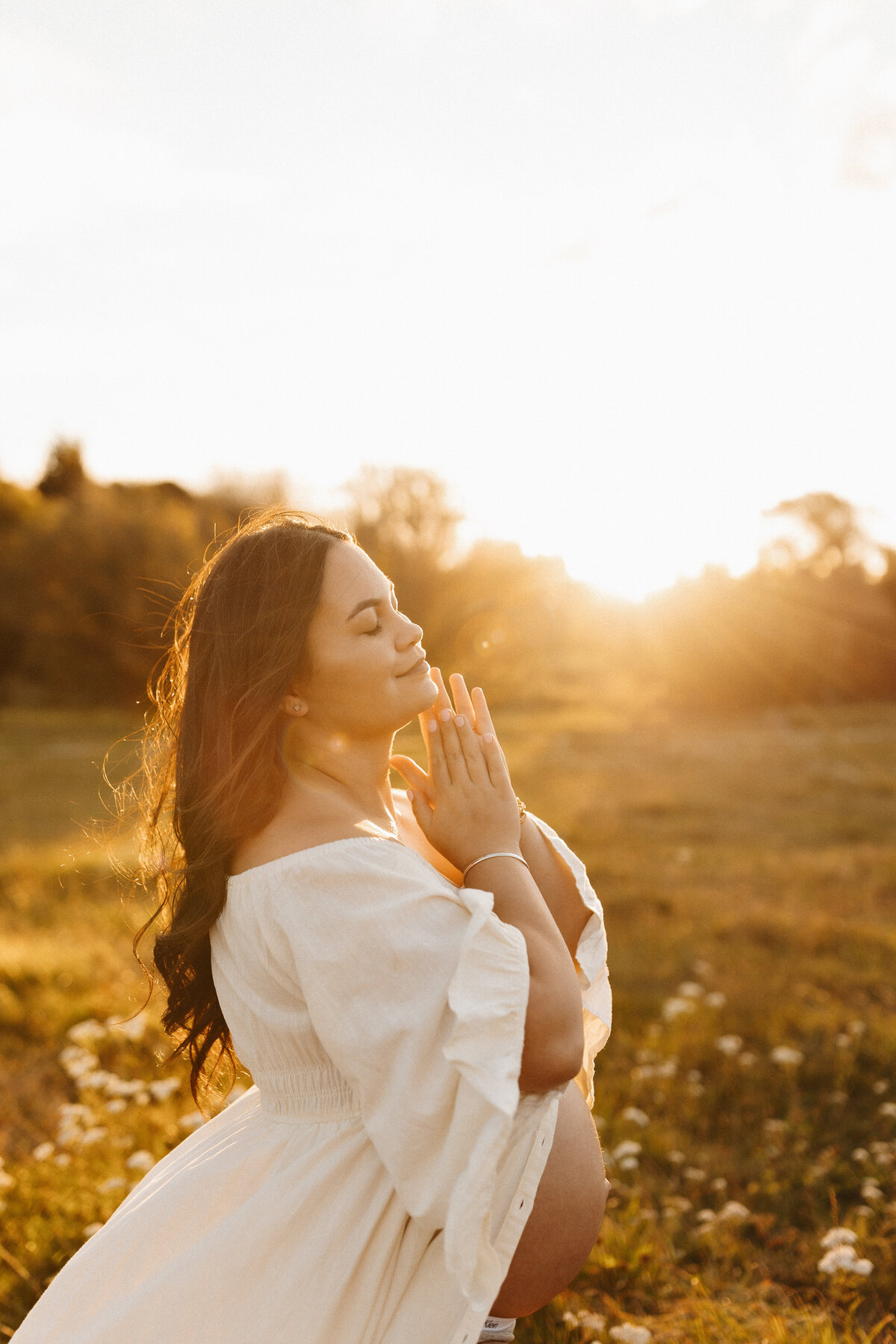 An expectant mother holds her hands to her face and relaxes into the sunlight wearing a white maternity dress during a photoshoot with lucy jane photography in christchurch