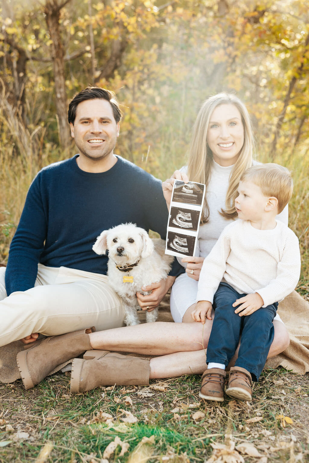 Two parents sitting in the grass with a small child and a dog as one holds up an ultrasound