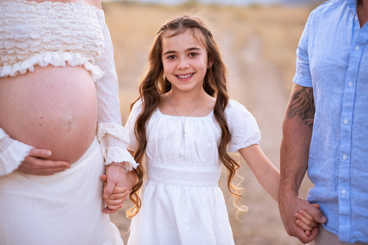 A little girl holds her mom and dads hands and smiles at the camera. Pregnant mom is on the left and dad on the riight, you can't see their faces.