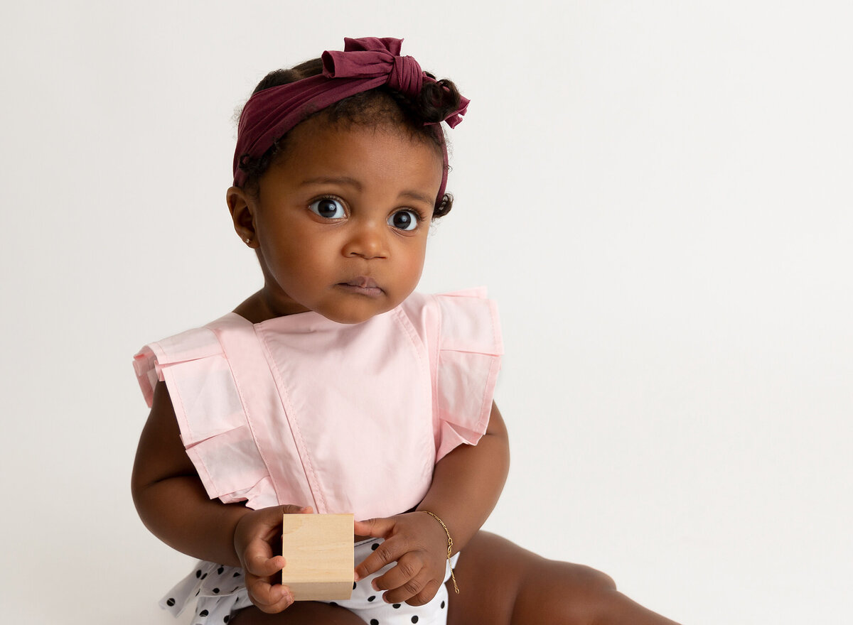 Baby girl is sitting for a 6-month baby milestone photoshoot. Baby is holding a wooden block and looking at the camera.