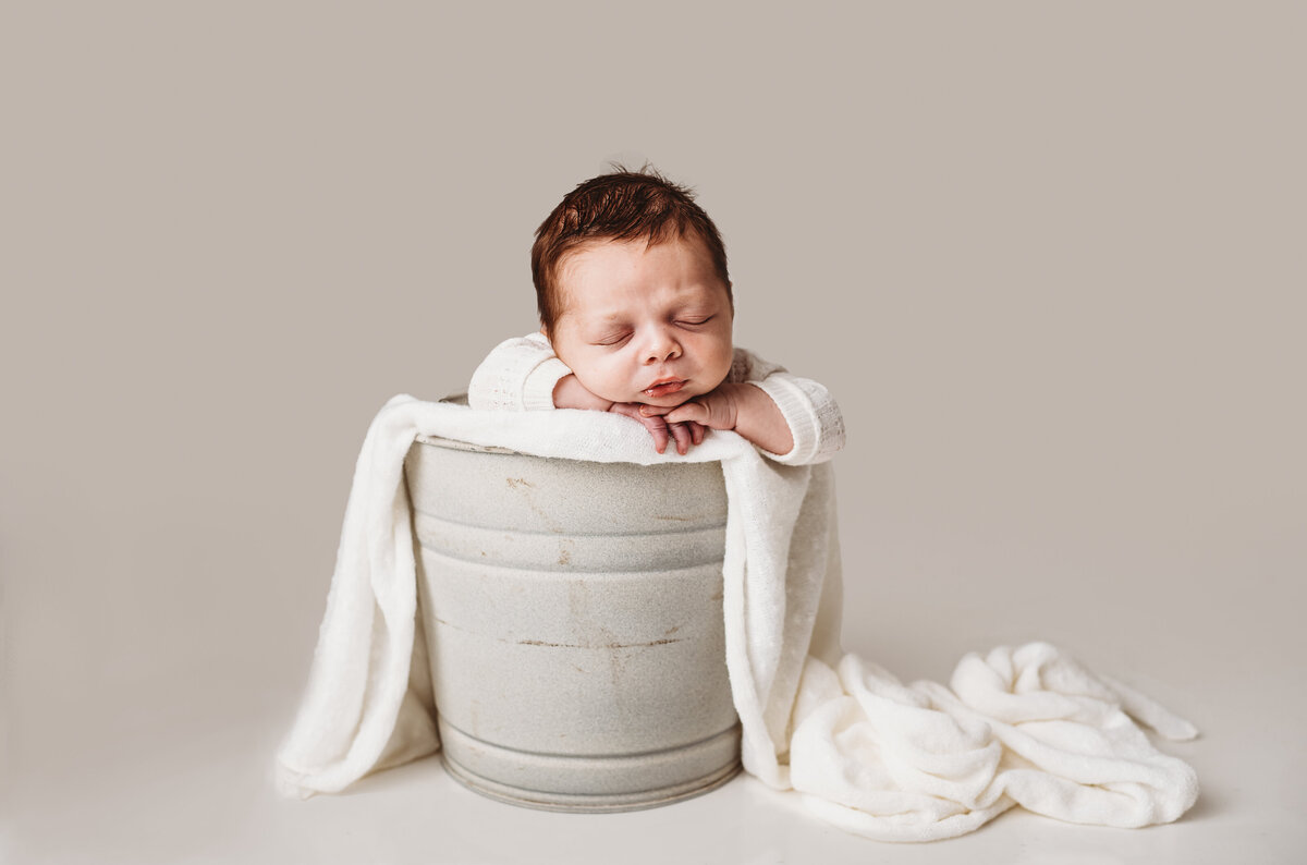 newborn photos with little baby in a white swaddle sitting in a bucket on a white background for an in studio session with Family photographers Maryland