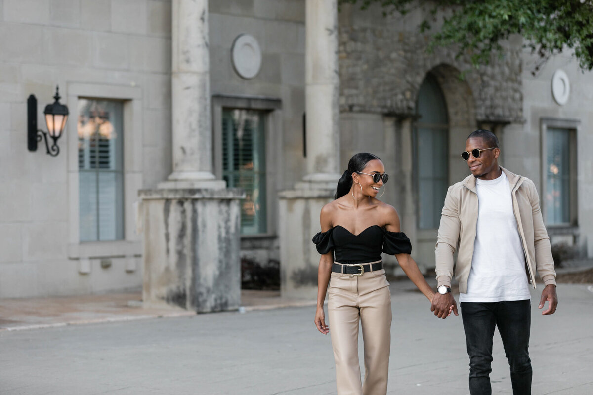 Stylishly dressed couple walking at Adriatica, holding hands and wearing sunglasses during their engagement session.