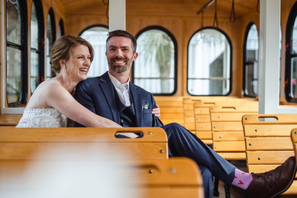 A bride and groom sit laughing on a vintage wooden trolley, their happiness contagious. The bride, in an elegant strapless lace gown, leans into the groom, who sports a sharp navy suit and a playful smile. His unique red socks peek out, adding a splash of color. The trolley's warm wooden interior and large windows frame the couple's joyous celebration.