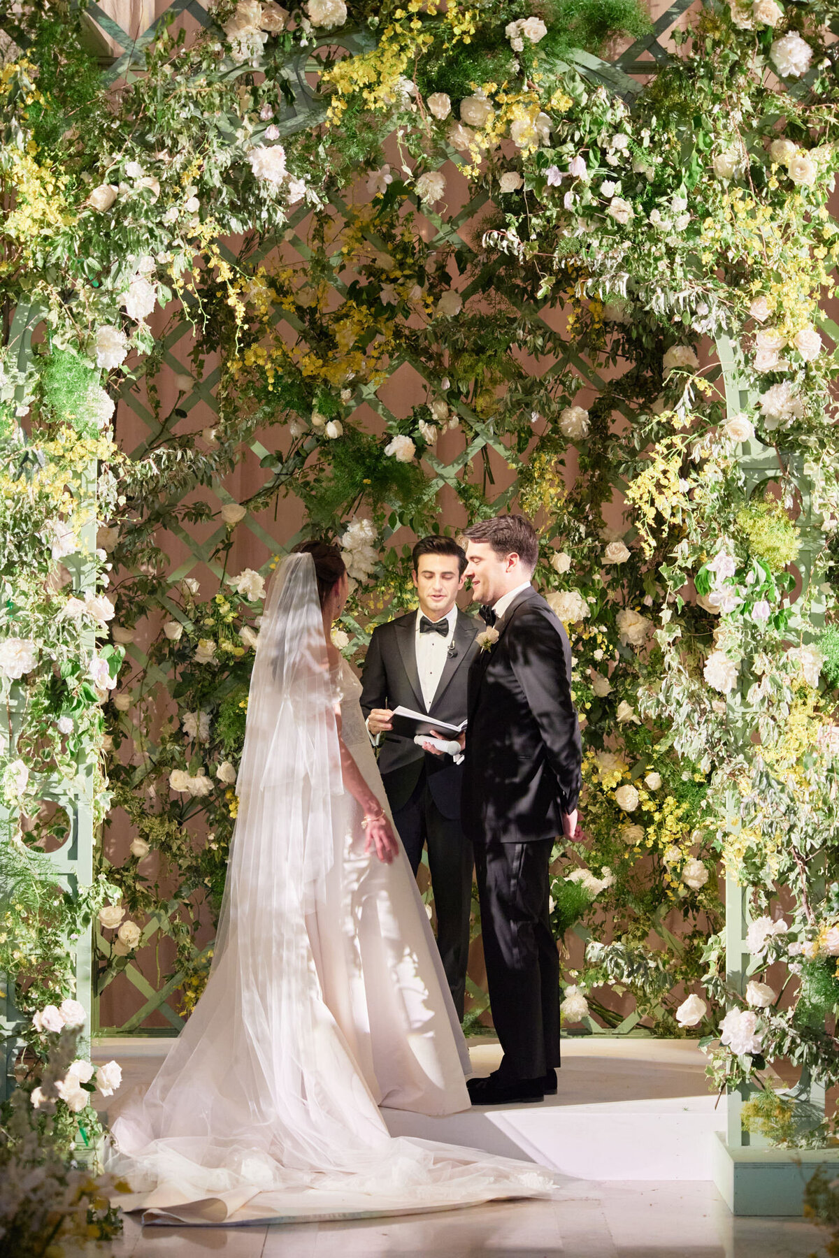 A bride and groom stand facing each other under a lush, floral archway adorned with green leaves and white and yellow flowers. The bride wears a long, flowing white gown and veil, while the groom is in a black suit. An officiant stands behind them, presiding over the ceremony.