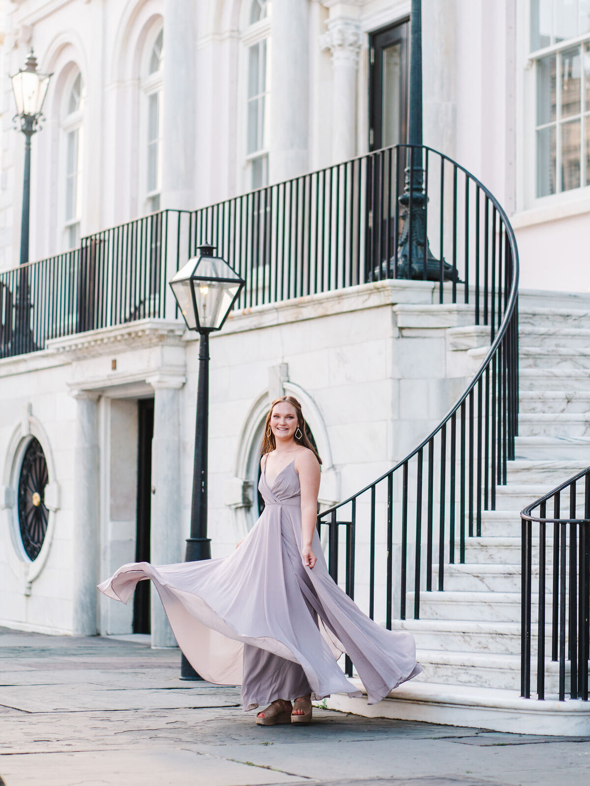 Senior girl in white dress and black hat leaning on railing of historic Charleston home