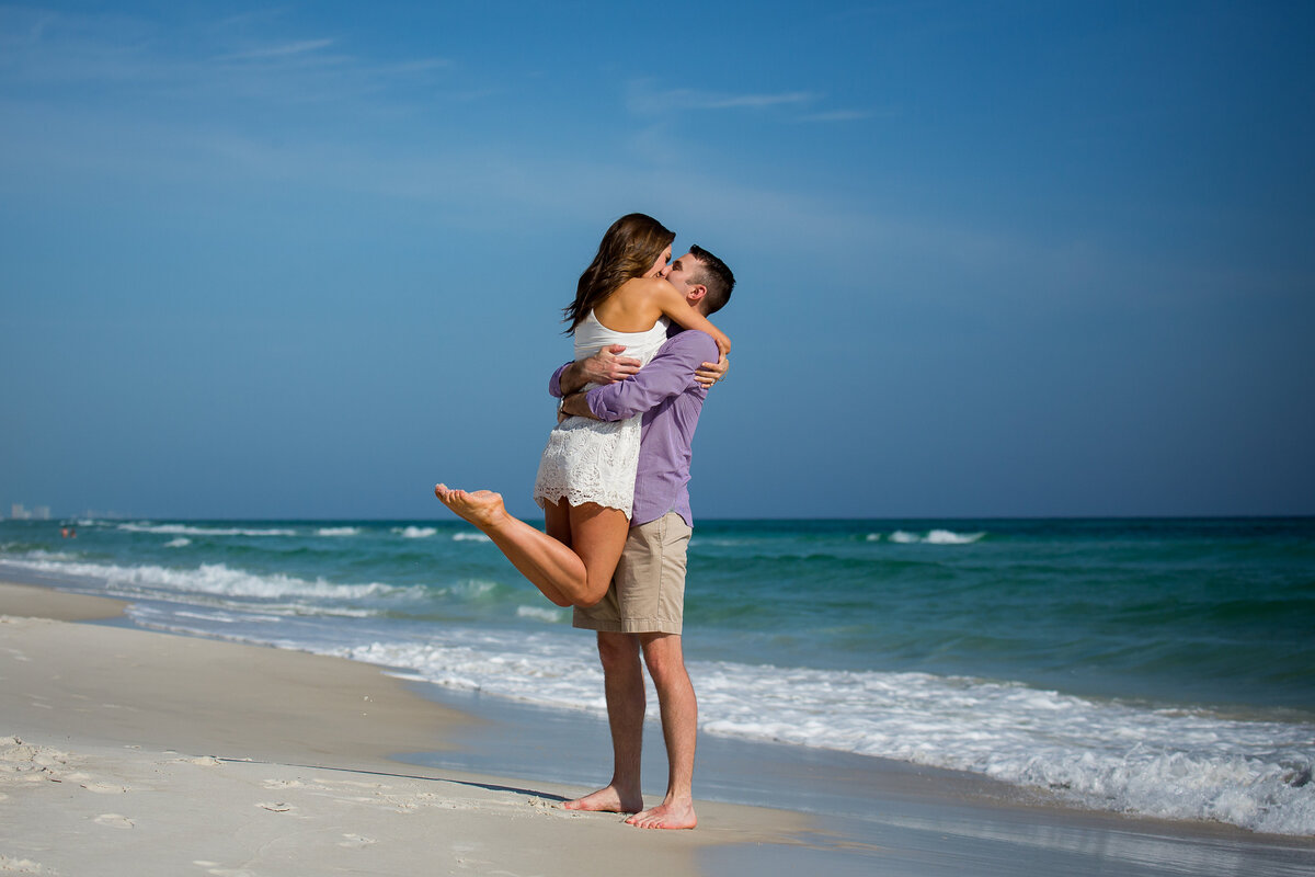 man holding woman up with her legs bent in front of the water at Rosemary Beach after she said yes to his surprise proposal.