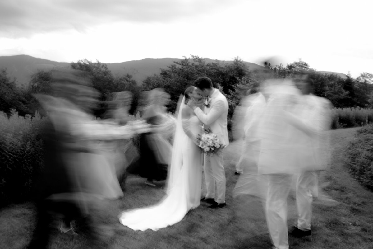 black and white photo of bride and groom kissing with bridal  party blurry as they walk behind