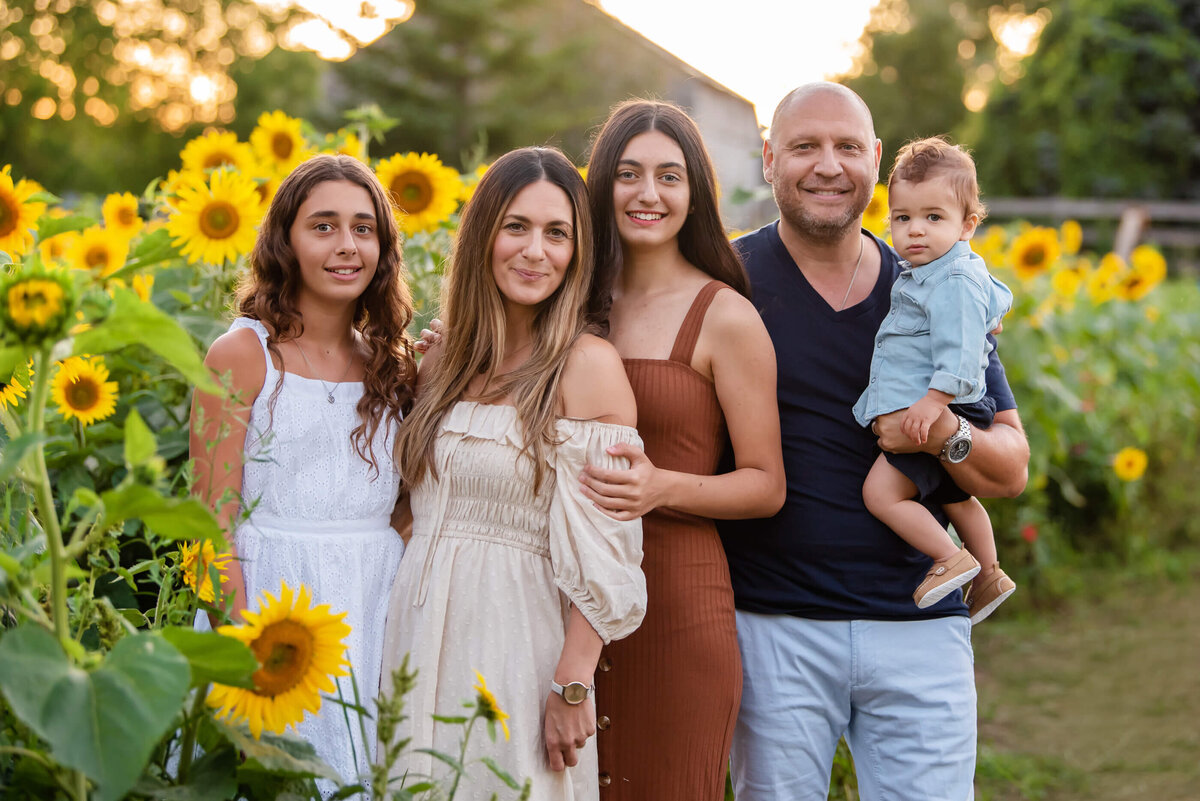 Up-close photo of a family of five with mom and dad, two teenage daughters and baby son in a sunflower field for their family photography session in Burlington, On