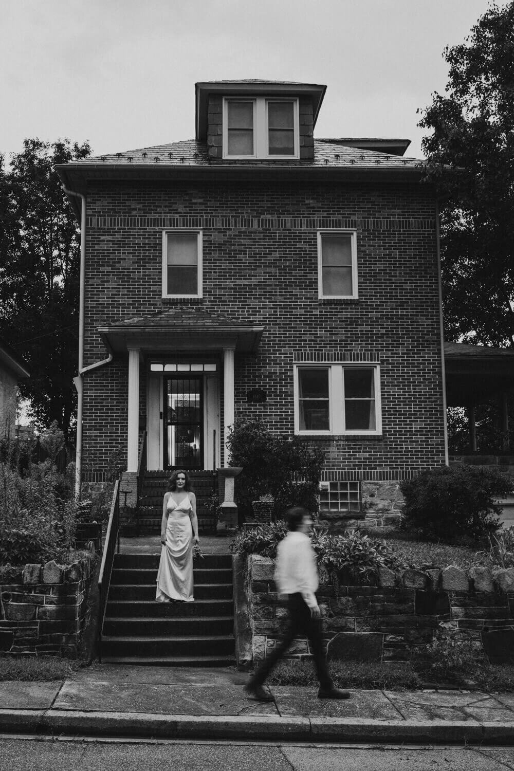 Black and white photo of guy walking in front of home while partner holds flowers in Baltimore