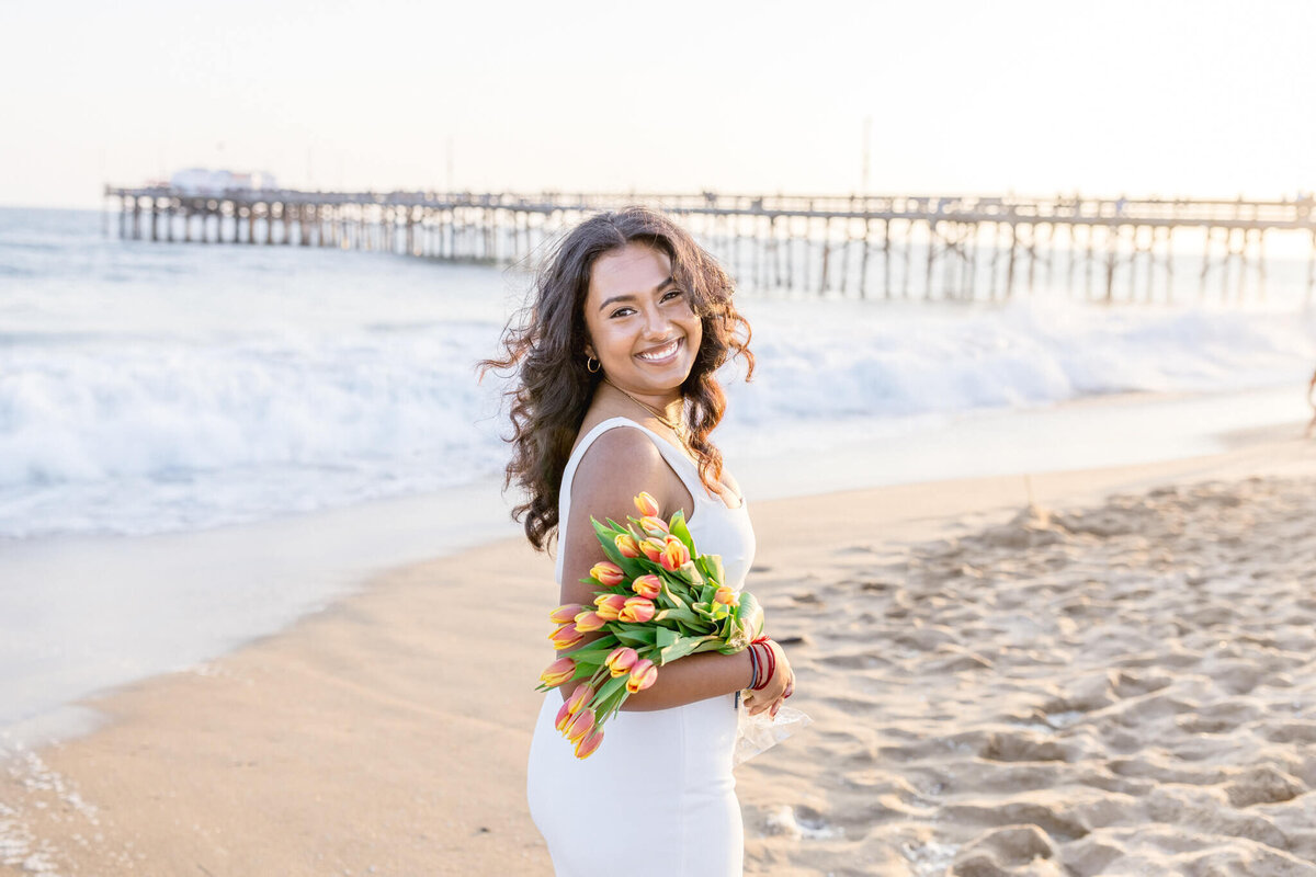 A beautiful young woman with long, wavy brown hair wearing a long white dress and holding a bouquet of yellow and pink flowers with green leaves in her right arm, looking over her right shoulder as she's standing on the sand of a beach, with the water and pier in the background.