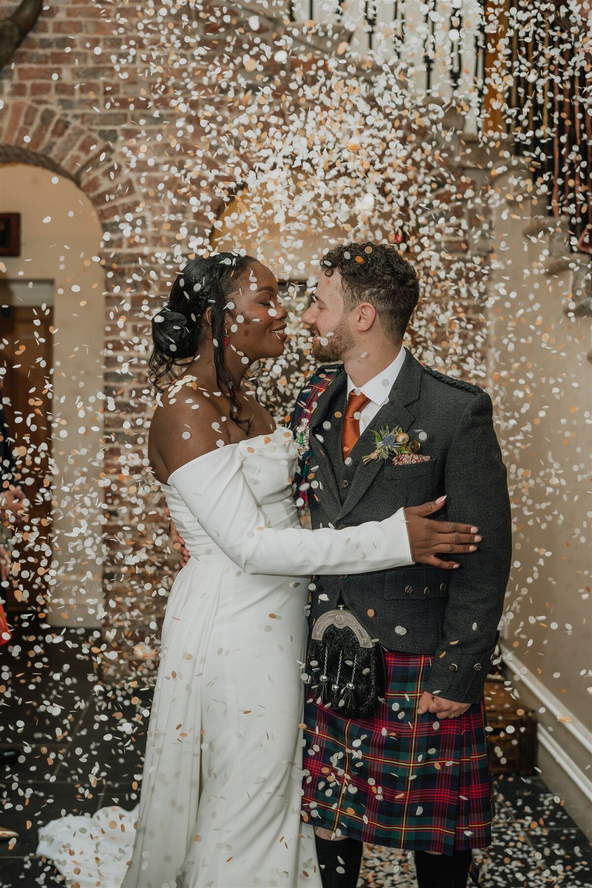 A bride and groom are photographed in the entry hall of Logie Country House as guests throw confetti from above them. The confetti falls like snow indoors and the couple smile at each other.