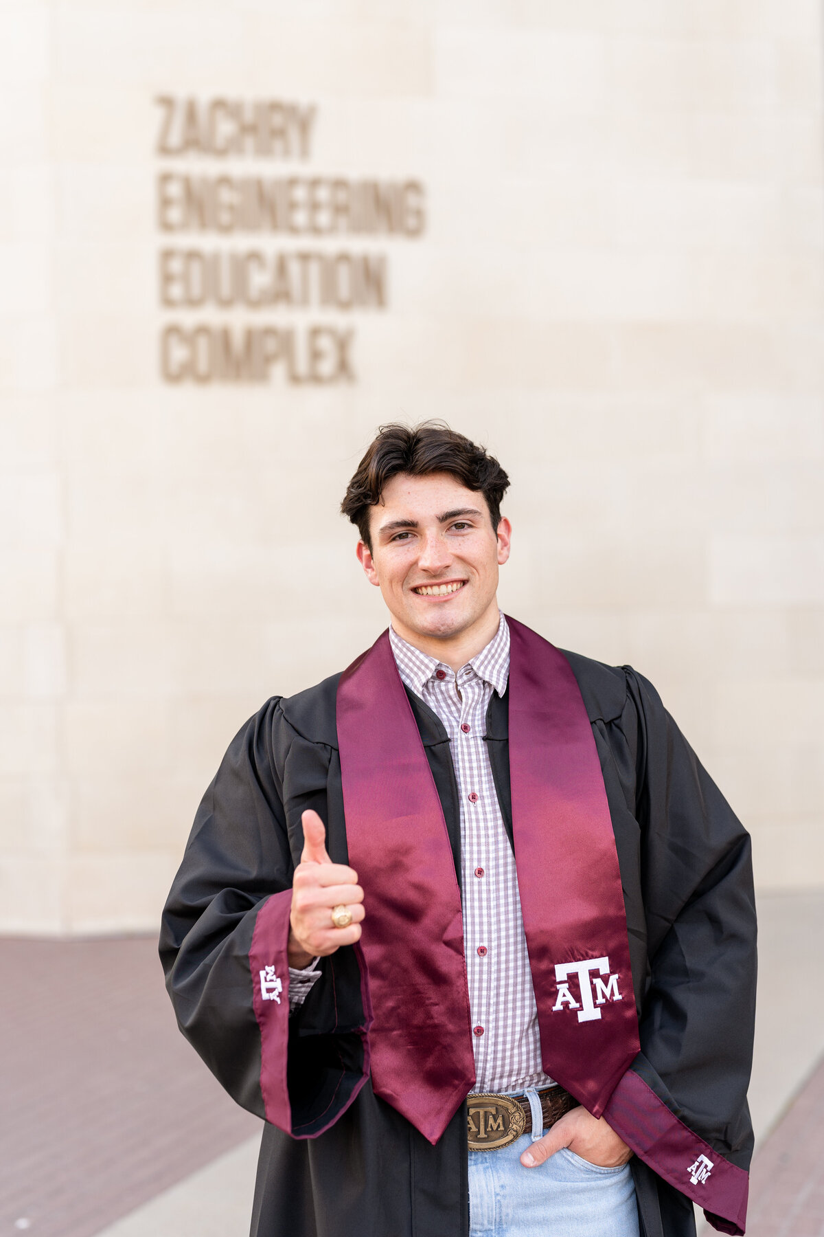 Texas A&M senior guy wearing gown and stole with hand in pocket and other thumbs up while smiling in front of Zachry Engineering Complex