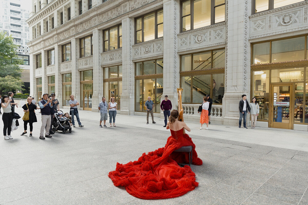 woman in long red gown is playing harp in the urban courtyard of the Wrigley Building, with numerous people listening as she plays.
