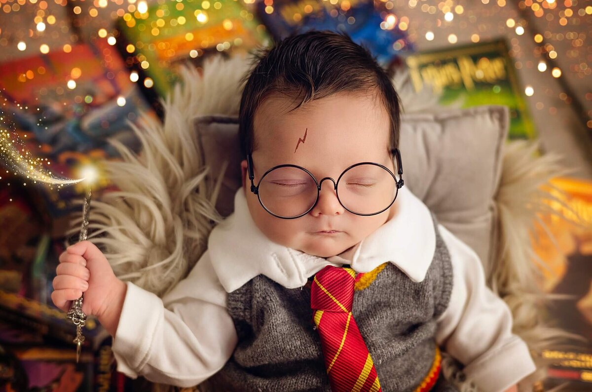 A baby, dressed in a grey vest and a red and yellow striped tie, sleeps soundly while clutching a silver wand. The baby is wearing round glasses and has a lightning bolt drawn on their forehead, reminiscent of the famous Harry Potter character. The baby is surrounded by books and a soft, white fuzzy surface, creating a whimsical and magical atmosphere. The background is blurred, with warm, golden lights that add to the enchanting scene. The image captures the innocence and wonder of childhood, combined with the magic and imagination of fantasy.  