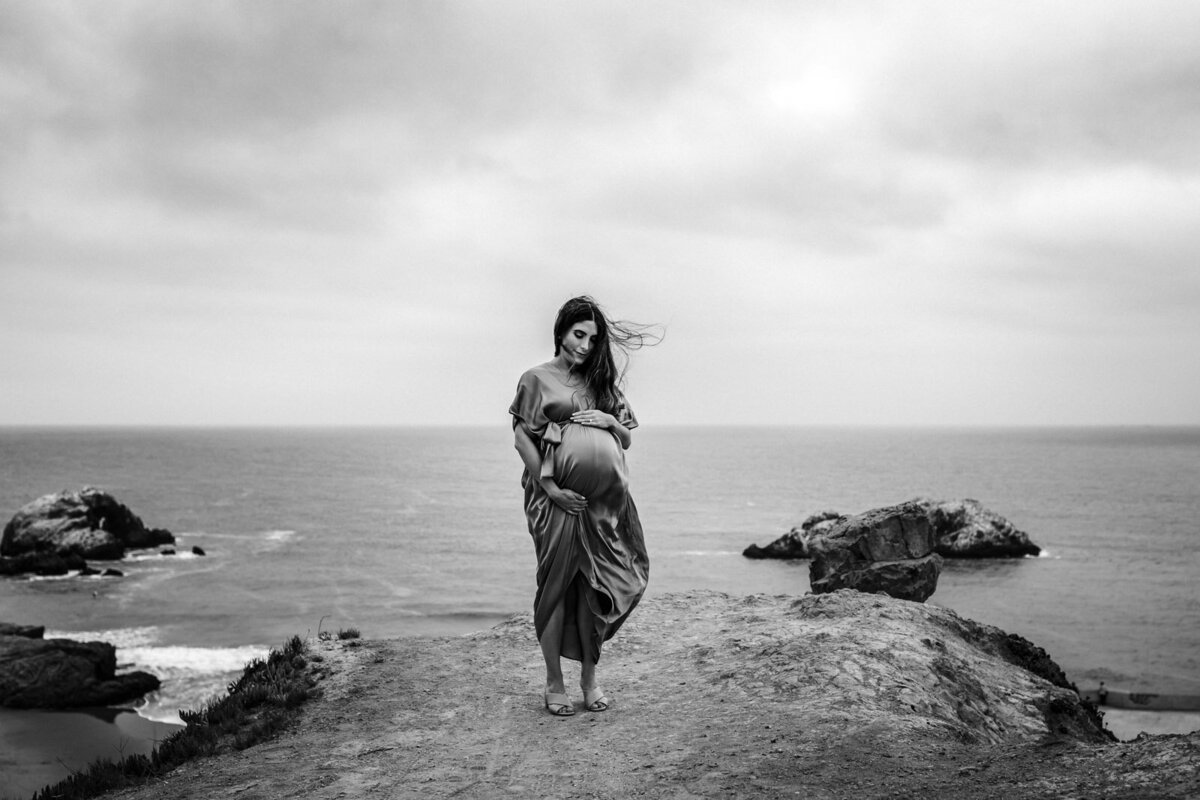 pregnant woman standing above the pacific ocean in San Francisco holding baby bump with hair and dress blowing in the wind